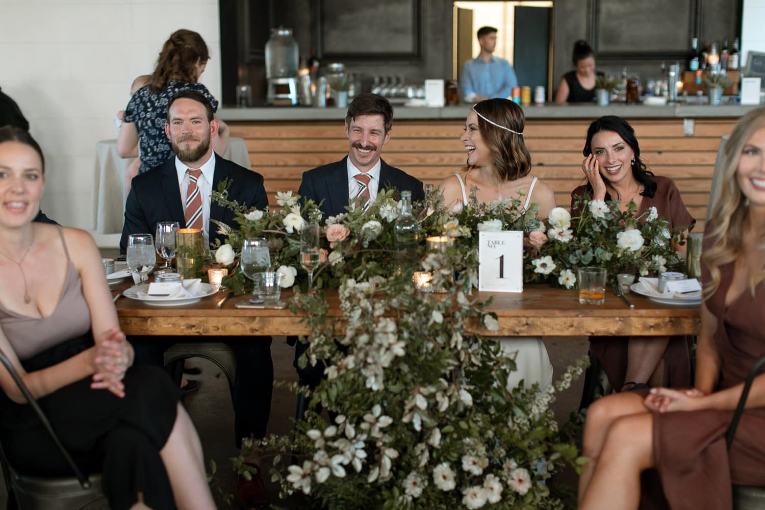 Bride and groom sitting at a table with white and greenery running down the centerpiece. There are candles. There are pops of peach in the roses. It looks very modern. Their friends are all laughing together.