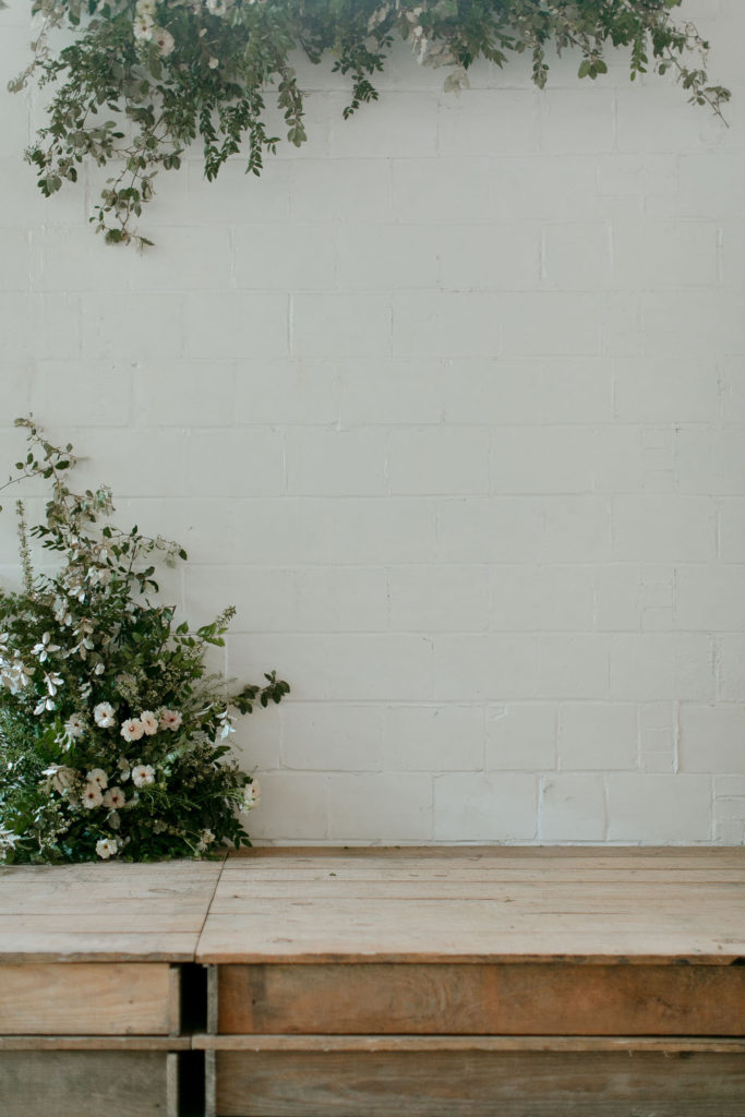 Wedding Ceremony decor with a white and greenery focus. The walls are white, the floor is concrete and there are candles on the ground. There are plants throughout the aisle and on the ceremony stage.