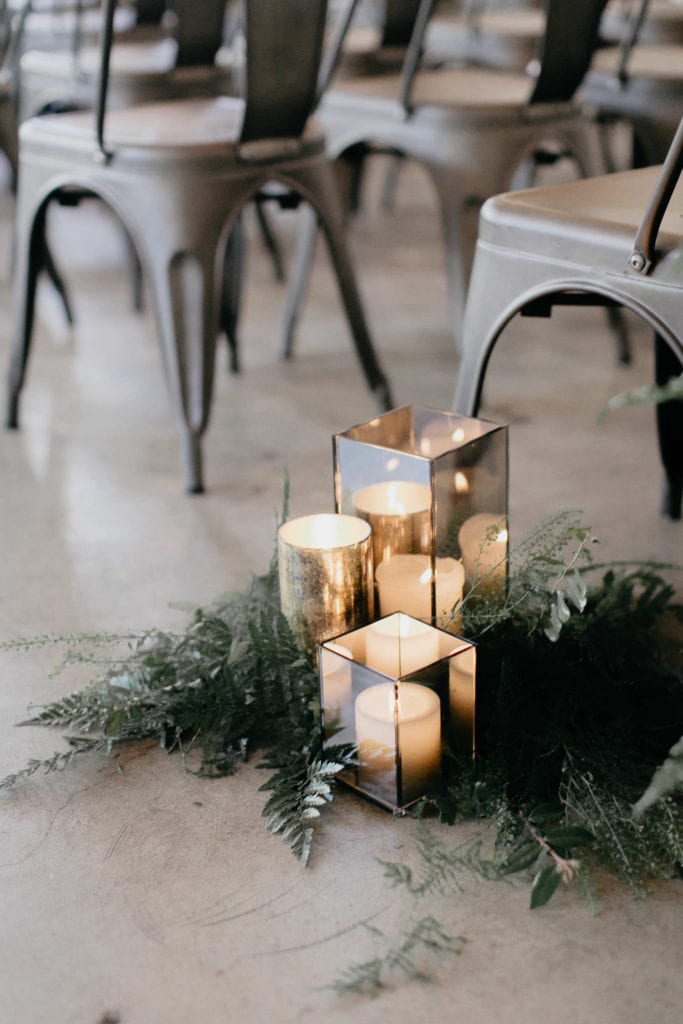 Wedding Ceremony decor with a white and greenery focus. The walls are white, the floor is concrete and there are candles on the ground. There are plants throughout the aisle and on the ceremony stage.