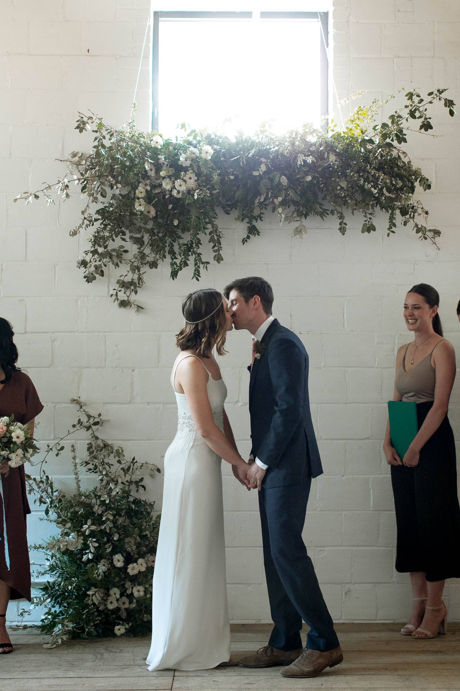 A couple kissing at their wedding ceremony which is decorated by white and greenery.