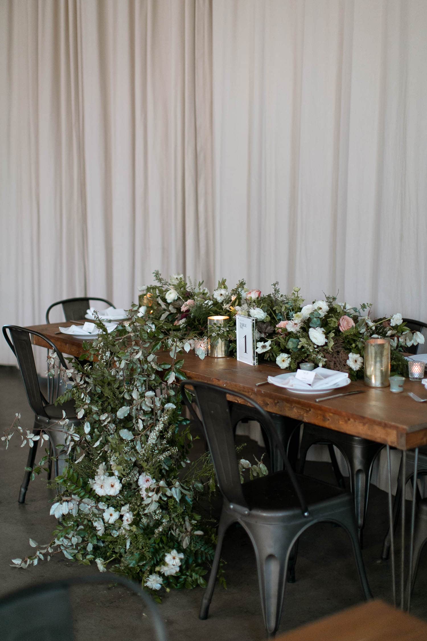 There are white walls, concrete floors, modern farm tables, greenery and candles on the tables. The sweetheart table has white flowers running down to the floor.