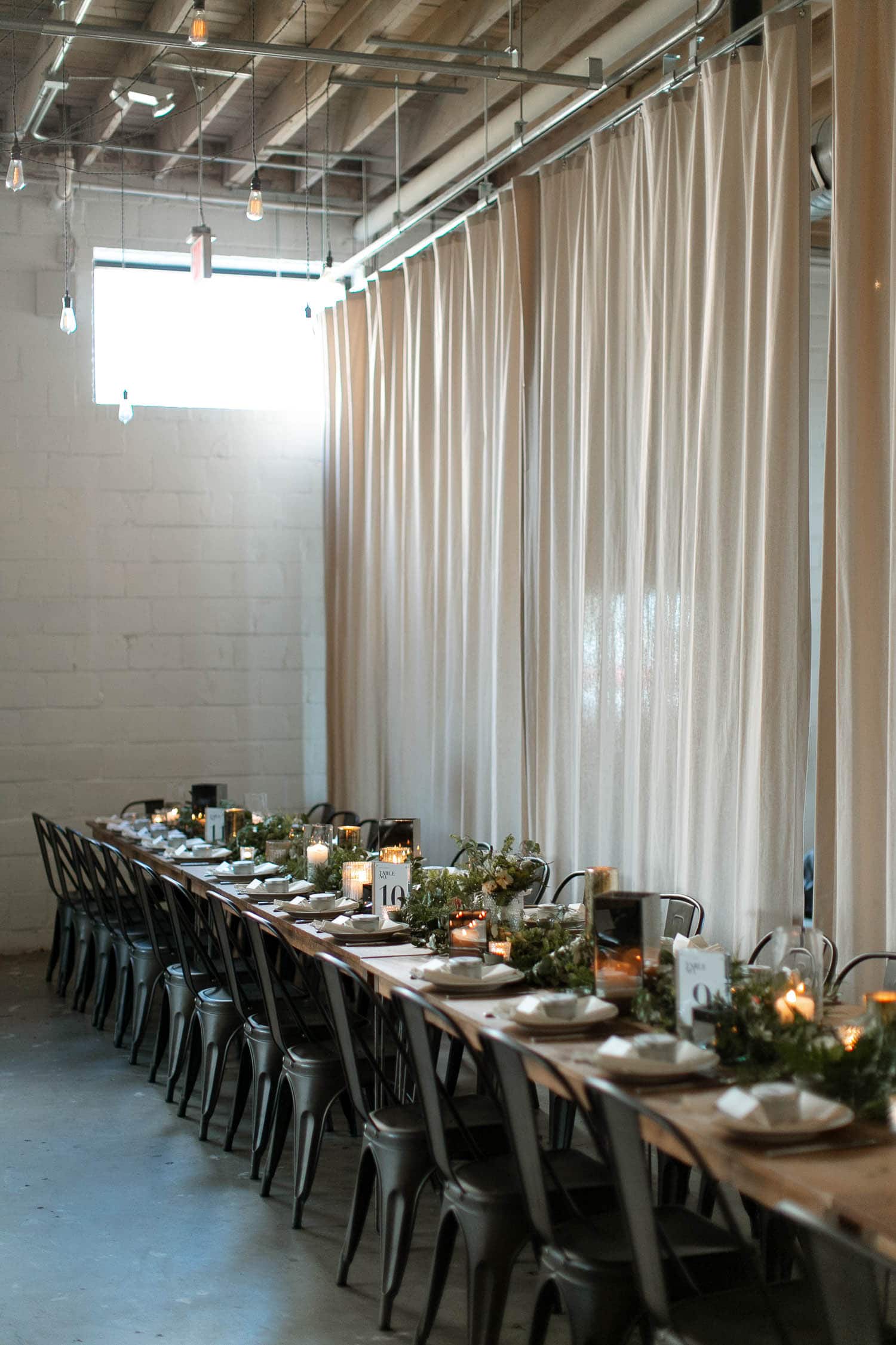 Wide angle of a wedding reception which has white and greenery decor all around. There are white walls, concrete floors, modern farm tables, greenery and candles on the tables. The sweetheart table has white flowers running down to the floor.
