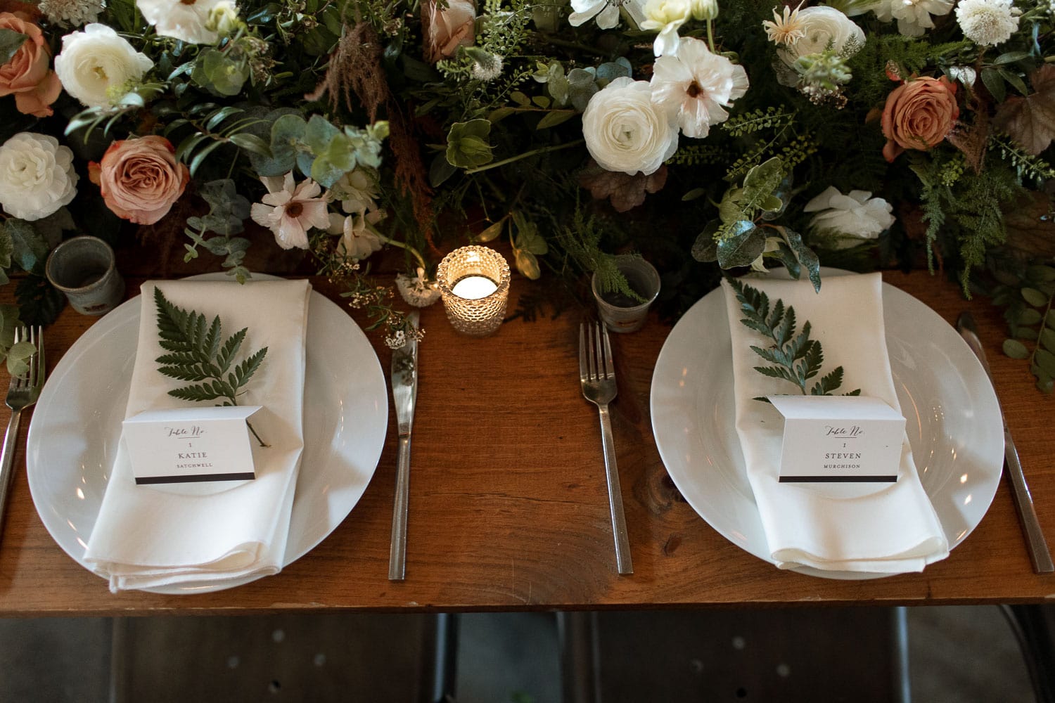Closeup of a table with white and greenery running down the centerpiece. There are pops of peach in the roses. It looks very modern.