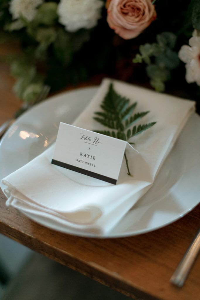Closeup of a table with white and greenery running down the centerpiece. There are candles. There are pops of peach in the roses. It looks very modern.