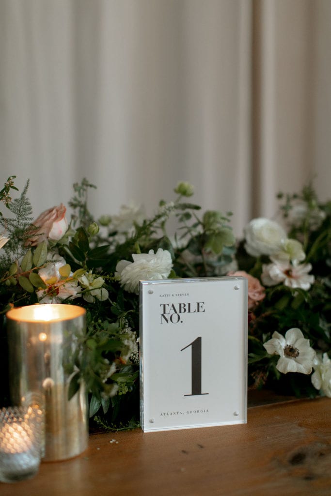 Closeup of a table with white and greenery running down the centerpiece. There are candles. There are pops of peach in the roses. It looks very modern.