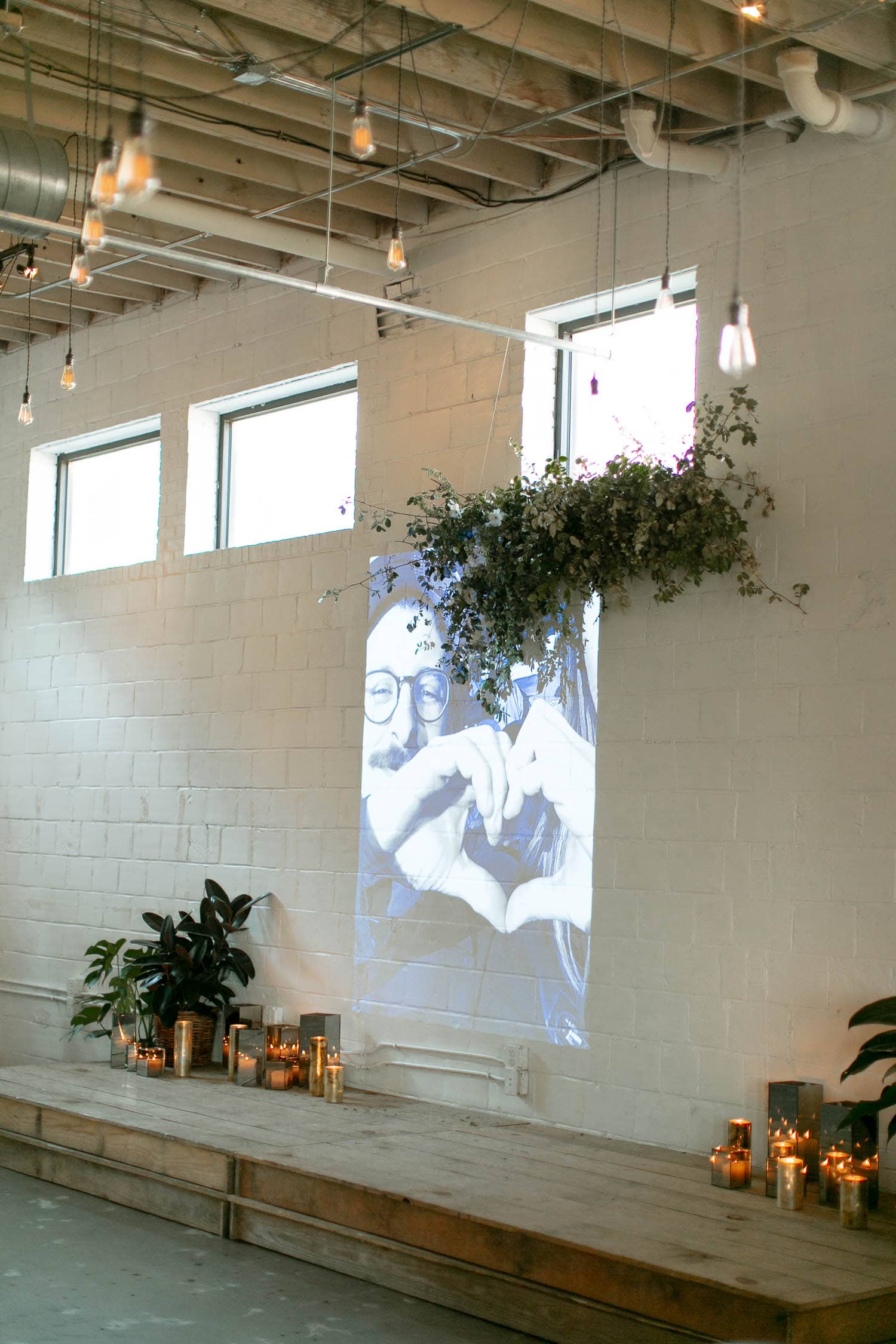A wedding ceremony stage has houseplants all around, adding to the white and greenery wedding theme. There is a projection of photos of the couple on the wall.